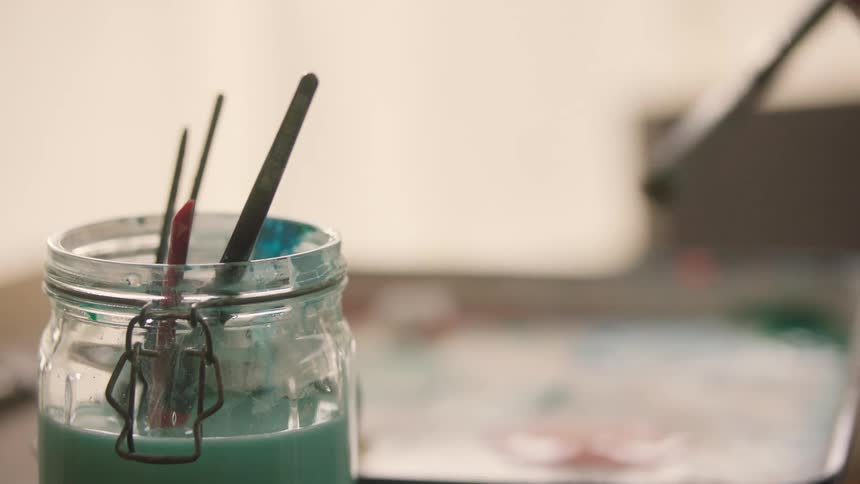 A hand adorned with a large gold ring and bracelet lifts a paintbrush from a jar of teal rinsing liquid then moves the brush toward a palette to mix into paint on a bright workspace