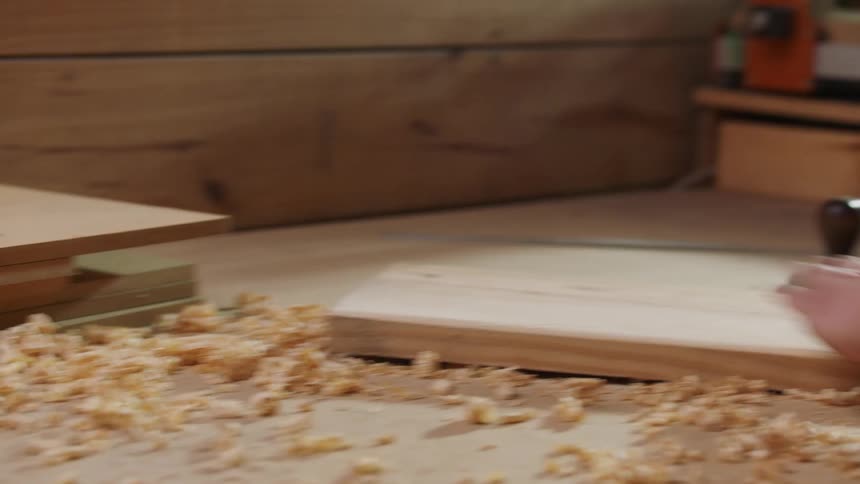A person in a plaid shirt sweeps wood shavings off a wooden board next to a hand plane