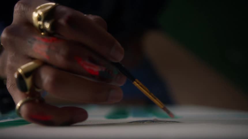 A closeup shot focuses on an artists hand which is marked with smudges of red and orange paint and adorned with several large gold rings as the person delicately holds a small brush to apply paint to a white surface