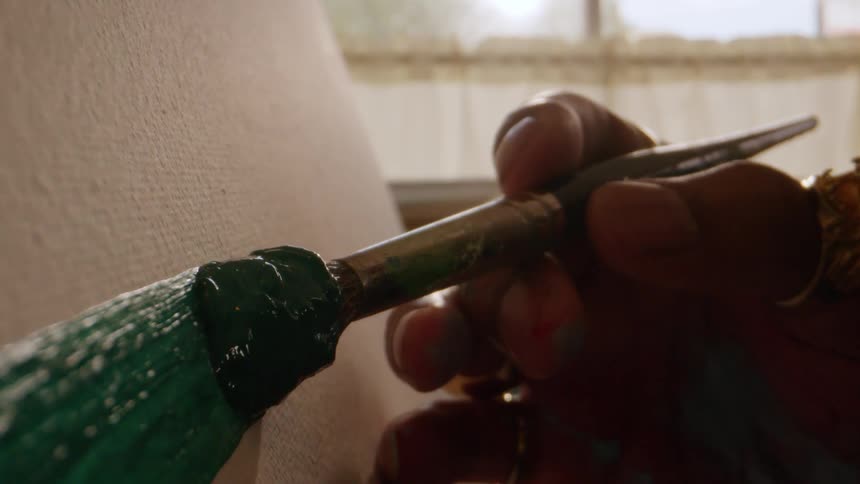 Brush spreading thick green paint on textured canvas with natural light in the background