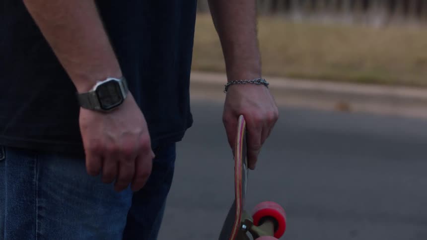 A man wearing cuffed blue jeans and canvas sneakers is shown first standing on a skateboard then picking it up by the deck and holding it by his side revealing the red wheels and a watch on his wrist