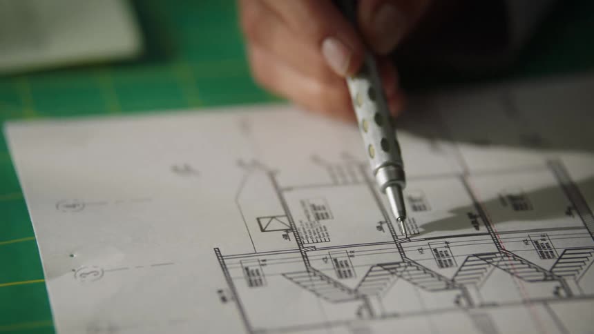 A closeup shot focuses on a persons hand using a mechanical pencil to carefully trace a line on a technical blueprint which rests on a green gridded mat