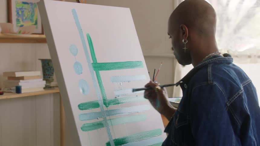 Artist painting a picture on canvas on his easel in his studio with plants and books on the shelves with art supplies