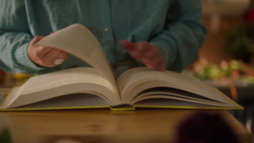 A person in a blue shirt opens a thick yellow book on a wooden table and flips through the pages