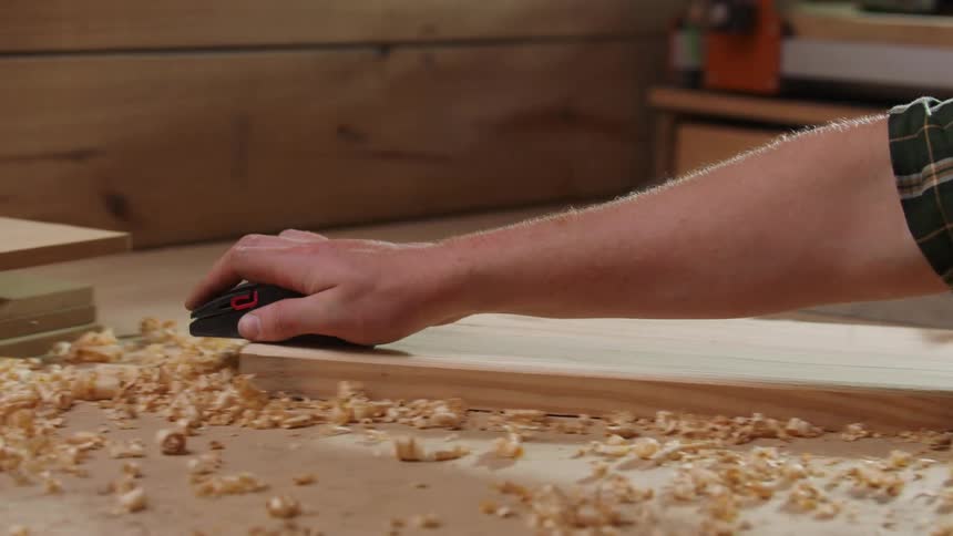 A view from the side shows a persons hands using a manual wood plane across a piece of lightcolored wood generating a large amount of wood shavings that cover the surface