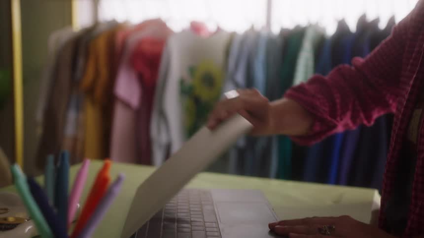 Person in red checkered shirt opening silver laptop in front of rack with colorful clothes