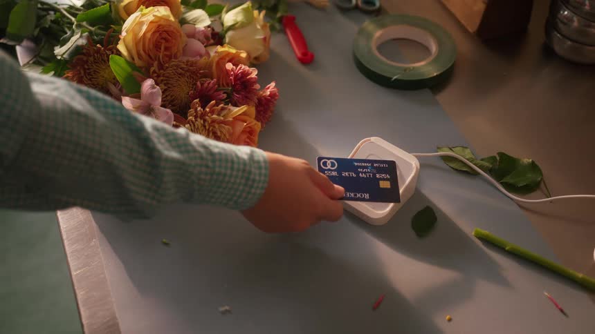 A person taps a credit card on a white reader sitting on a counter next to a bouquet of flowers