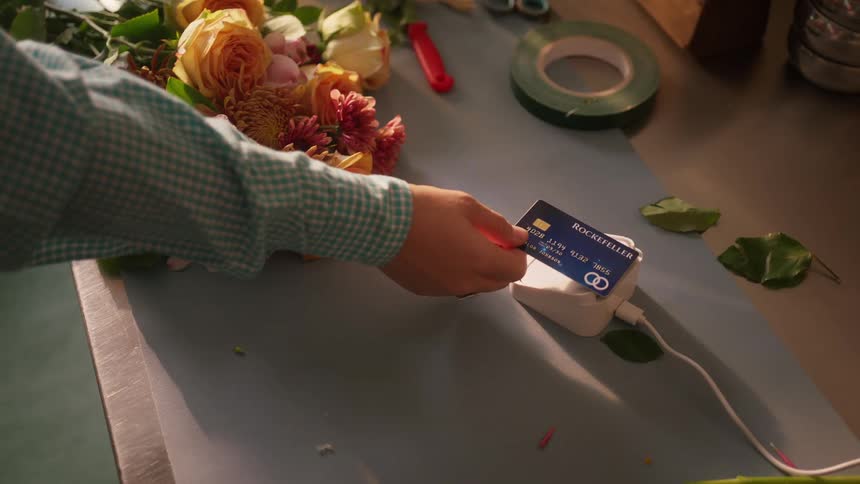 The video shows a floral arrangement featuring orange and pink roses and mums on a work surface a hand quickly swipes a blue and white credit card through a small white card reader next to the bouquet