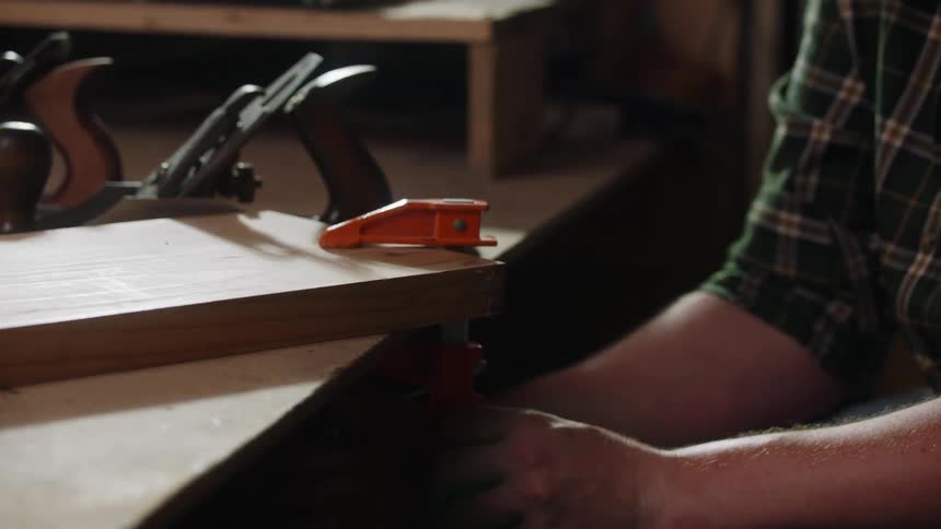 A person in a plaid shirt secures a wooden board to a workbench using a clamp