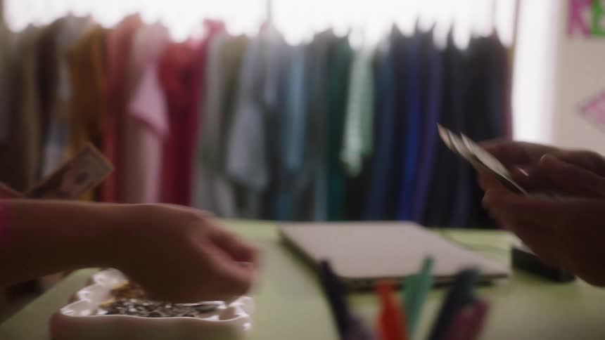 A person in a pink shirt hands cash to someone across a counter with clothing racks in the background