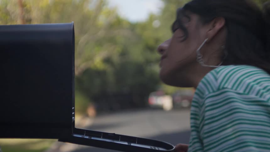 A young woman opens a black metal mailbox at the edge of a suburban street she peers inside checking for mail on a sunny day