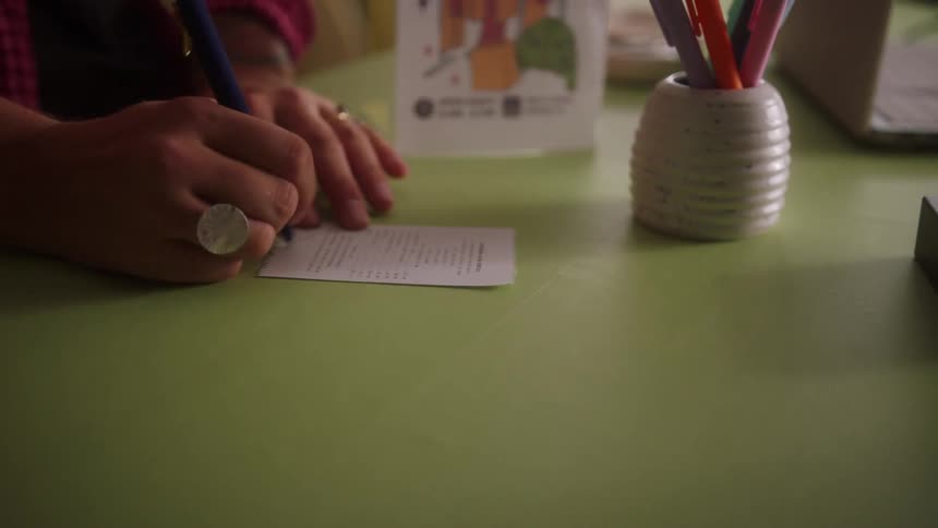 Hand writing on small paper on green table surrounded by stationery and electronics