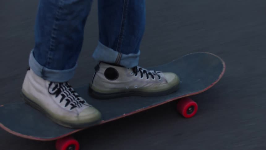 A person rides a short dark skateboard with red wheels across a paved surface