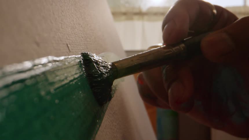 Hand holding paintbrush applies thick green paint on surface in naturally lit setting