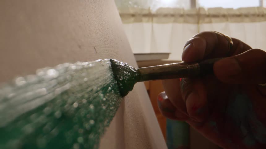 The video is a very closeup shot showing a paintbrush held by a darkskinned hand sweeping a thick line of deep green paint onto a canvas or textured wall the background is brightly lit suggesting daylight near a window