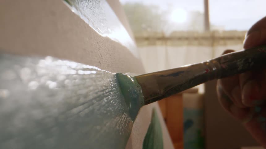 An extreme closeup focuses on a paintbrush held by a hand which is actively spreading a thick layer of light bluegreen paint onto the textured white surface of a canvas in a brightly lit room