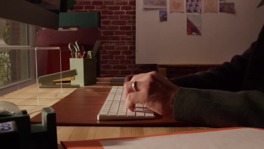 Person typing on white keyboard at wooden desk in creative workspace with brick wall background