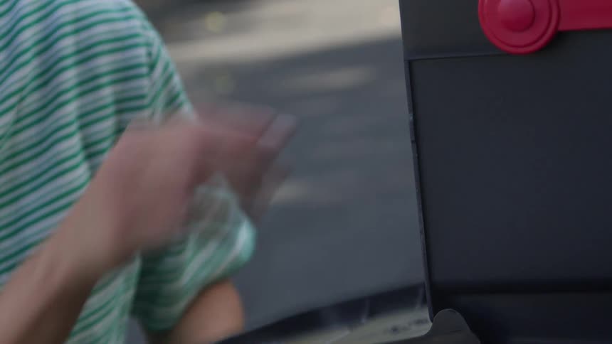 A closeup shot focuses on a woman wearing a green and white striped shirt as she reaches toward a dark mailbox open the small door and retrieve a handful of correspondence