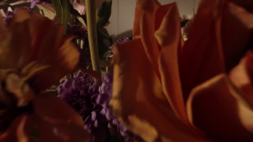 A closeup shows orange and purple flowers arranged in a dimly lit indoor space