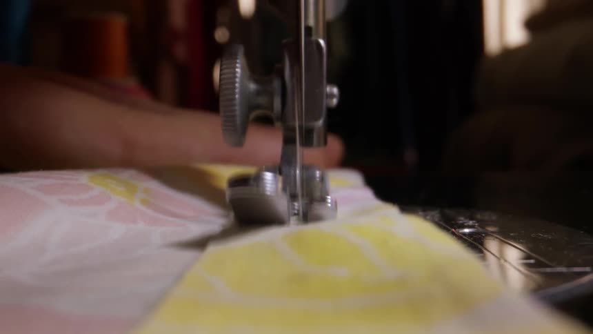 A dramatic closeup focuses on the chrome presser foot and needle bar of a sewing machine showing the needle rapidly stitching a piece of yellow and white patterned fabric as it is fed through the mechanism
