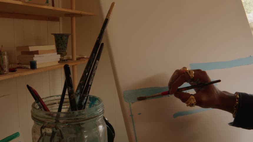 A closeup view focuses on an artists hand adorned with several gold rings painting a thick horizontal stripe of light blue paint onto a large white canvas while a jar filled with various paintbrushes sits in the foreground and wooden shelves