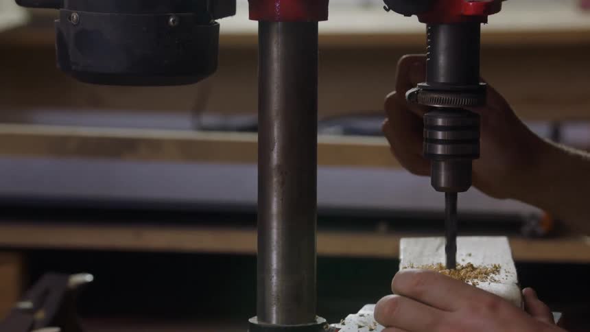 The video shows a person turning on an industriallooking drill press by flipping a switch on the red housing the machines drill bit descends and quickly bores a hole through a small piece of wood creating a pile of sawdust