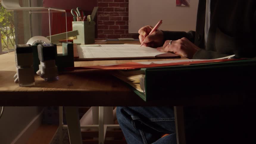 A person sits at a cluttered wooden desk and carefully writes in a desk calendar with warm light illuminating their hands and the workspace