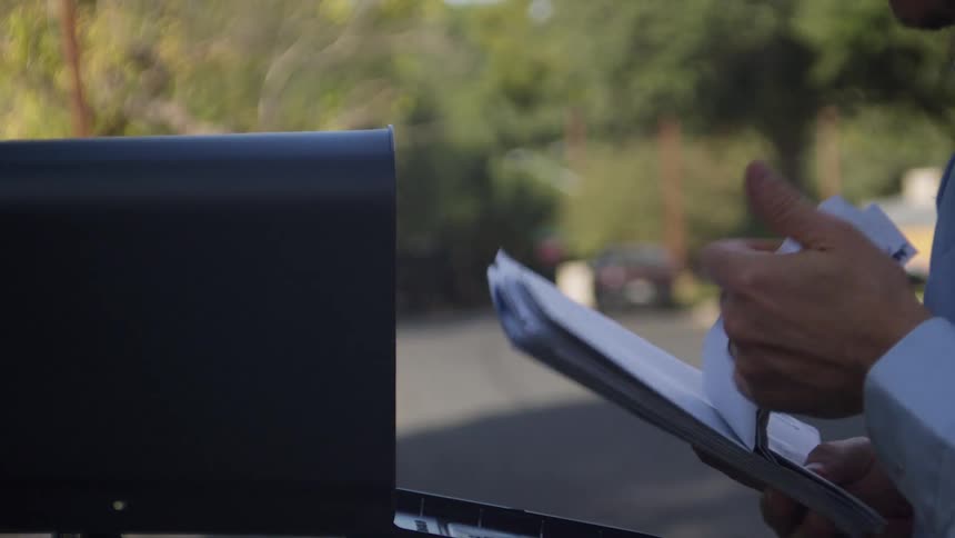 A mailman wearing a blue tshirt and carrying a black bag opens a wallmounted black mailbox to insert a stack of letters pausing briefly to verify the mail before sliding it inside