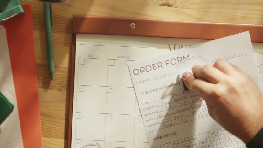 An overhead view focuses on a wooden desk where a persons hand is actively flipping through and moving a stack of completed forms and stamping a new one revealing a large monthly calendar pad beneath the work area