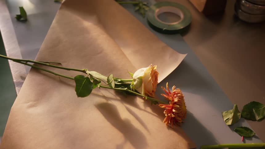 A person wearing a striped shirt is seen carefully arranging and gathering several cut flowers including yellow roses and an orange flower onto a large sheet of brown wrapping paper on a work surface preparatory to creating a bouquet