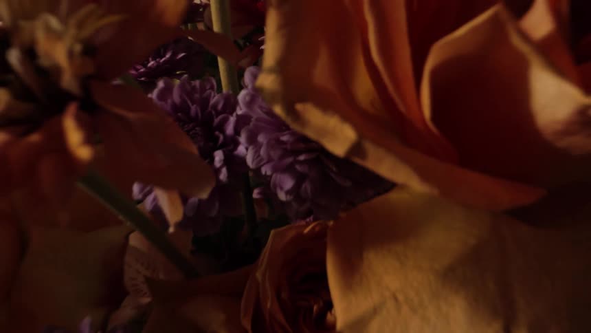 A richly colored arrangement of fresh flowers including orange roses and purple chrysanthemums resting in a vase next to a woven basket in soft indoor light