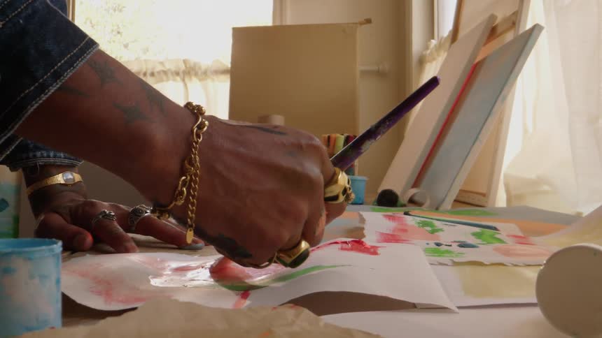 A persons hand holding a paintbrush picking up a color from a table and then applying it to the paper