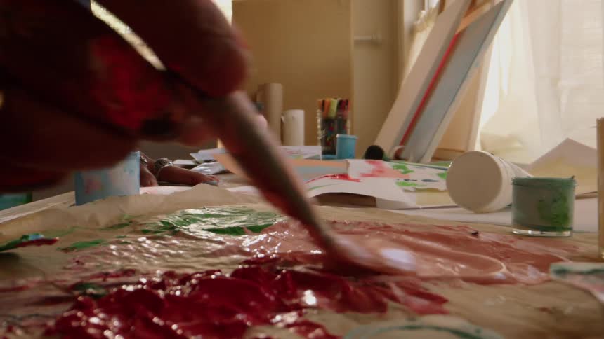 An artists hands move from a work on paper to a messy palette mixing vibrant paints with a brush in a sunlit and creative studio workspace