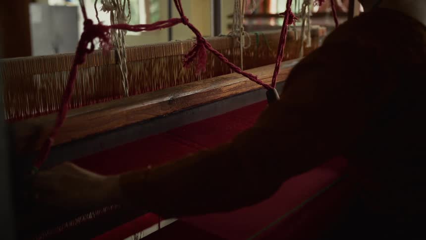 A persons arm operates the wooden mechanism of a traditional loom where a section of tightly stretched orange warp threads is visible and a bright section of vibrant pink weft fabric is being woven