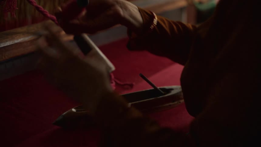 The video is a closeup dimly lit shot focusing on a persons hands operating a weaving loom the hands are skillfully manipulating red thread and passing a shuttle across the rich red fabric being woven