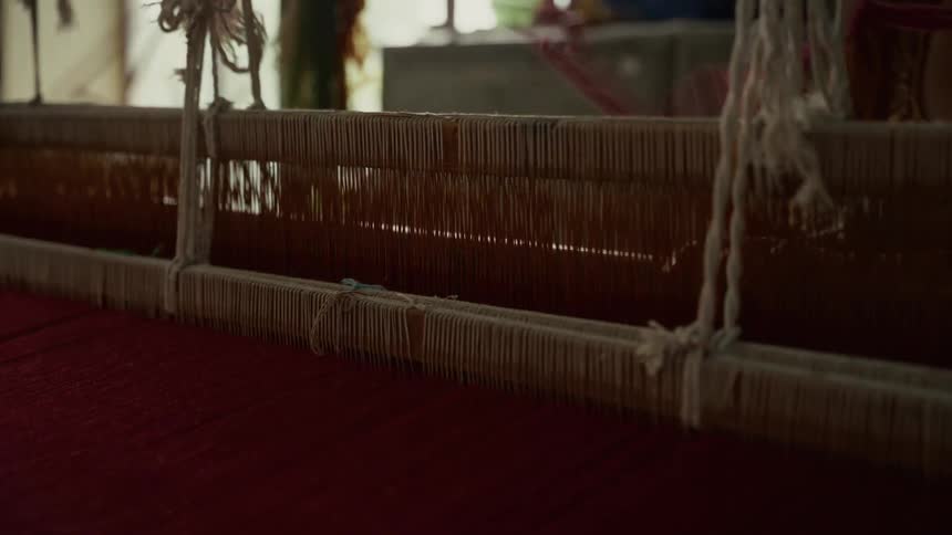 A closeup shot captures the complex rhythmic movement of the pale warp threads being raised and lowered on a traditional handloom actively weaving a deep red fabric