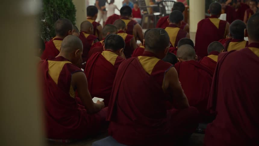 A large group of buddhist monks in red and yellow robes sit together on the floor of a temple courtyard some quietly reading from books