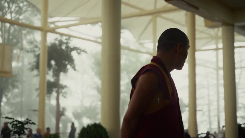 A man wearing the traditional reddishmaroon and yellow robes of a buddhist monk is seen from the side walking past large circular interior columns toward an expansive mistshrouded outdoor area where trees and a group of people are visible