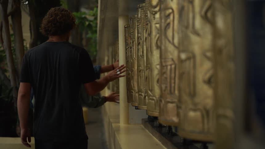 Three persons viewed from behind walks along a wall of large golden tibetan prayer wheels and uses their hand to spin the cylinders as a spiritual gesture