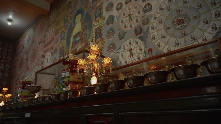 The video shows an interior view of a buddhist temple or shrine featuring a long shelf holding rows of offering bowls the wall behind the shelf is decorated with ornate and colorful thangkastyle murals and mandalas