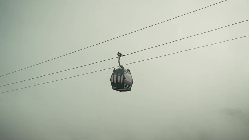 Empty cable car moves across the wire under a cloudy sky with a forested mountain in the distance