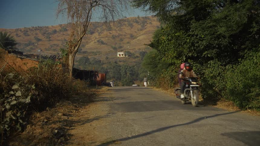 Person riding motorcycle on rural road flanked by dry vegetation and hills in the background