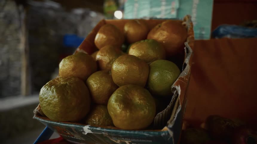 A weathered cardboard box filled with fresh oranges sits on display at a dimly lit market stall their rinds glowing under the soft light