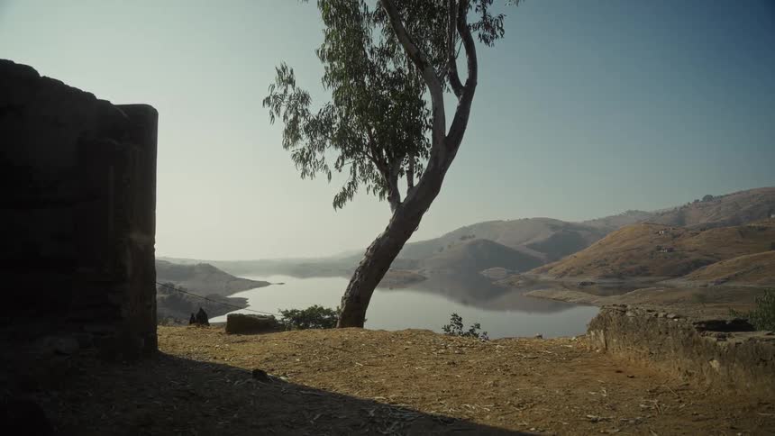 A tree stands beside a building wall at midday with a lake visible in the background along with hills and mountains
