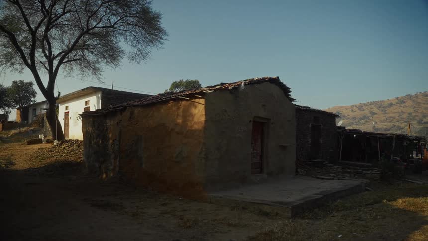 A static wide shot captures the exterior of a simple singlestory rural house made of earthenwalls and a clay tile roof situated in the foreground with another white building and dry scrubcovered hills behind it under a bright clear sky