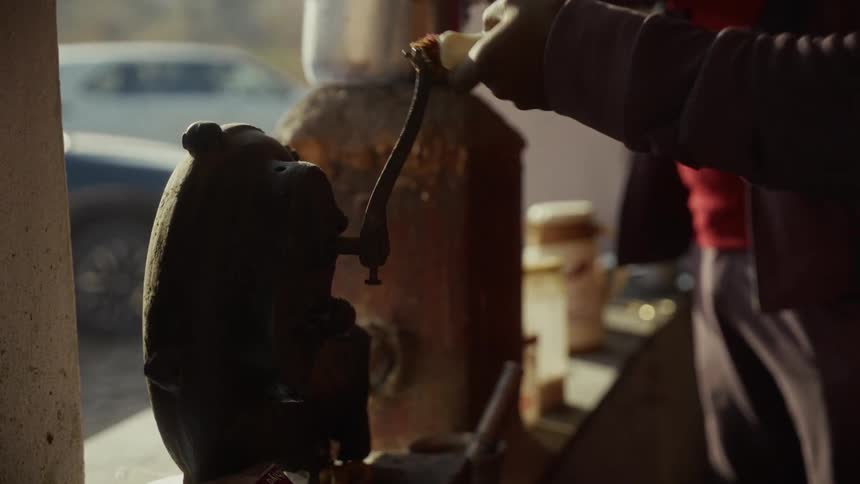 A person turns the crank of a dark cast iron machine in a sunlit outdoor setting
