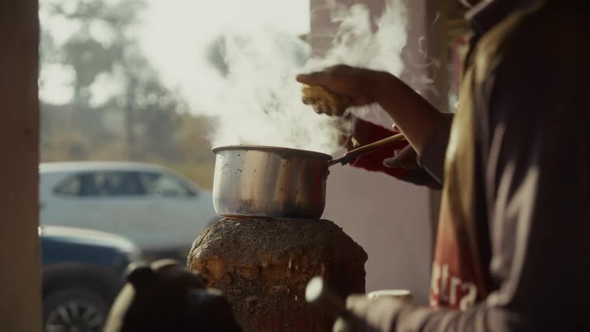 The video captures a steaming metal pot sitting on an outdoor heat source likely preparing chai or another hot drink a persons hands are visible using a ladle to stir and pour the contents of the pot