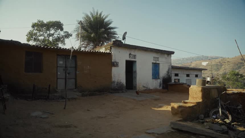 Two small singlestory homes one with a mudcolored exterior and the other whitewashed stand next to each other on a dusty unpaved patch of groundthe scene suggests a semirural or hilly location with a palm tree visible between the houses and dry