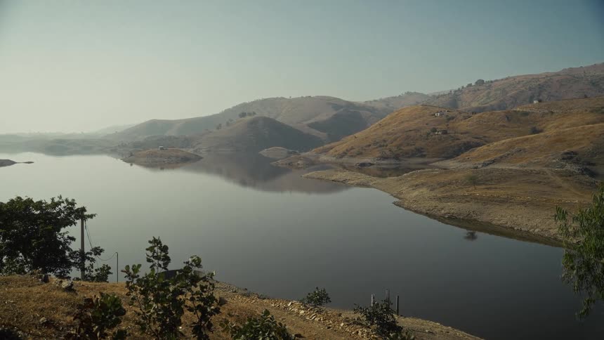 A tranquil lake sits nestled between arid rolling hills its glassy surface reflecting the pale hazy sky in a vast and quiet landscape