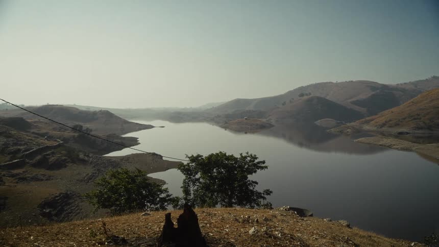 A calm body of water possibly a reservoir stretches into the distance reflecting the hazy sky and the surrounding landscapethe water is framed by sloping hills covered in dry lightbrown vegetation with a few green trees visible in the foreground